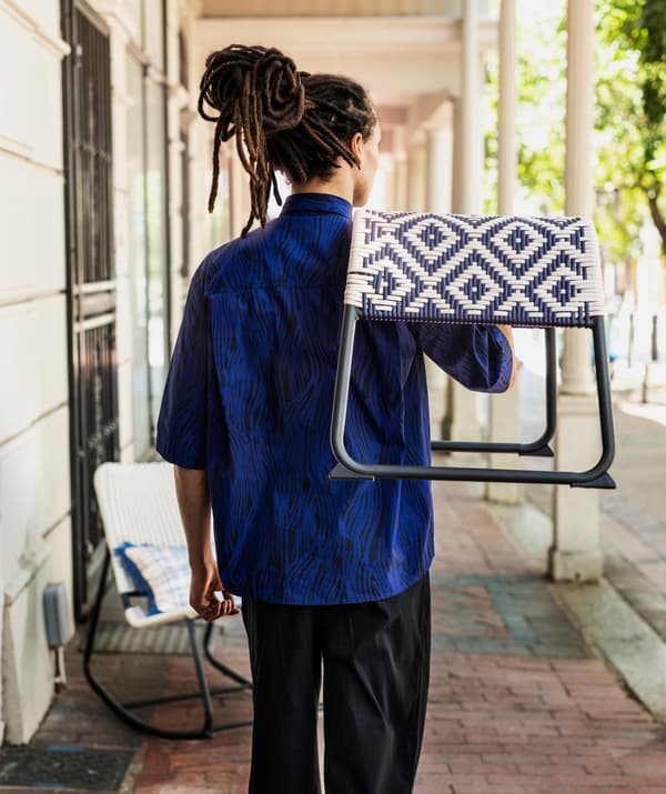 A man in a blue shirt carries a blue and white patterned ÖVERALLT footstool on his shoulder outdoors.