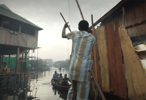 A man in a colourful suit punting on a boat through a floating wooden shanty town.