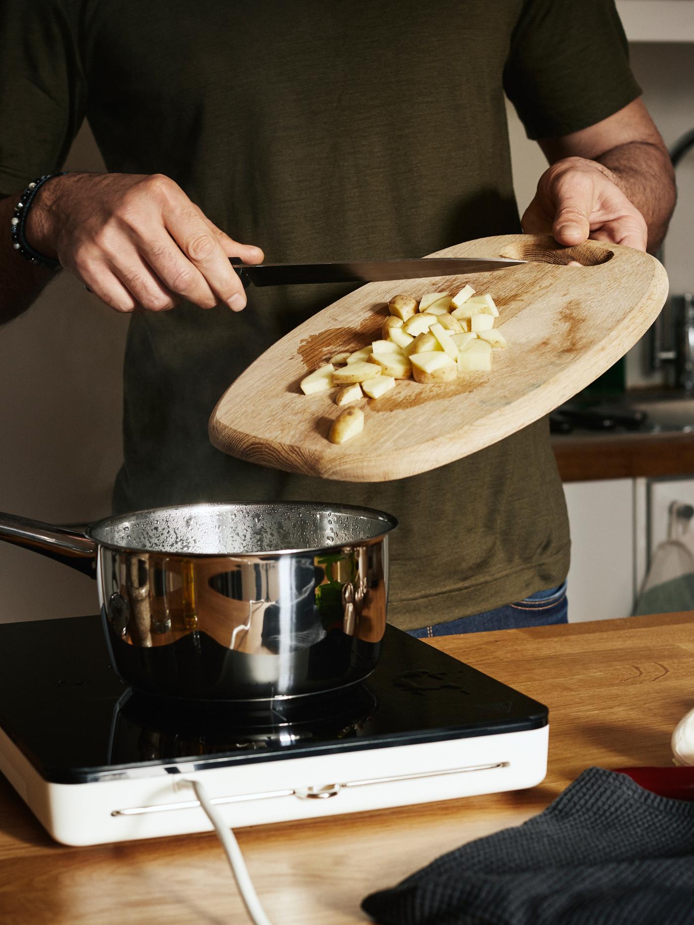 A man in a green t-shirt scrapes diced potato from a wooden chopping board into a pan on a TILLREDA portable induction cooktop.