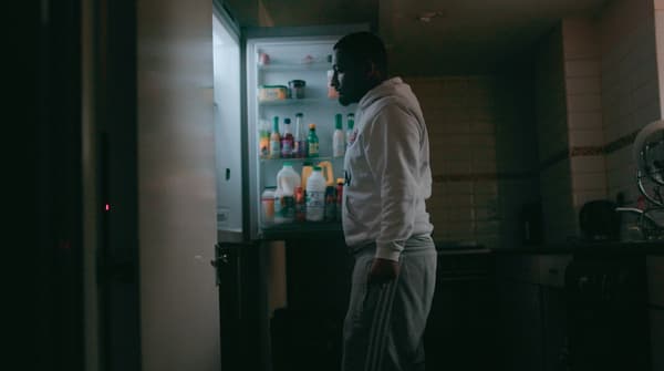 A man in a white hoodie and grey jogging pants is standing in a dark room, looking inside a fridge.