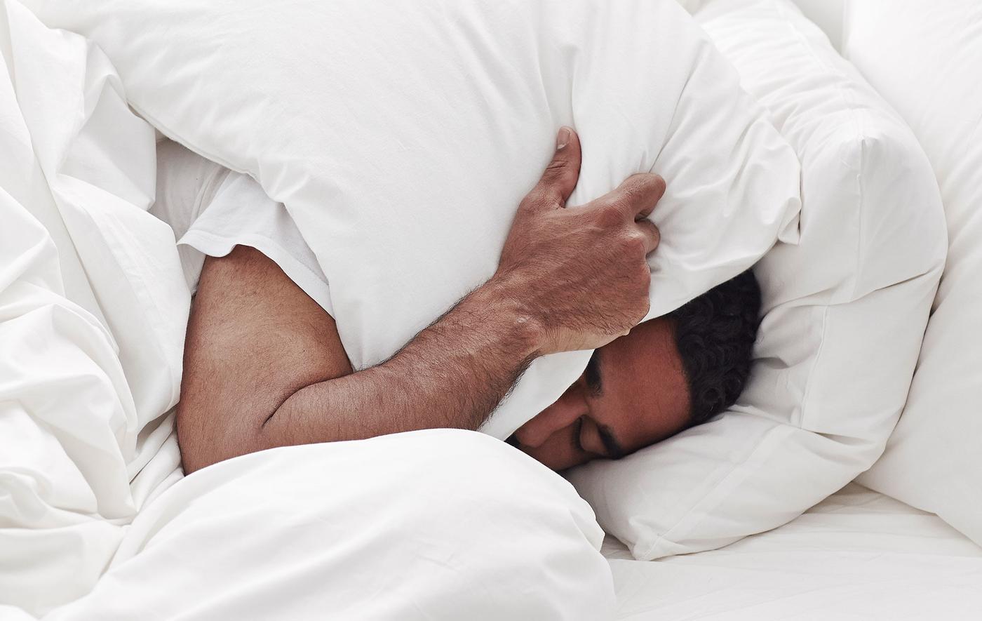 A man in bed covering his ears with a pillow.
