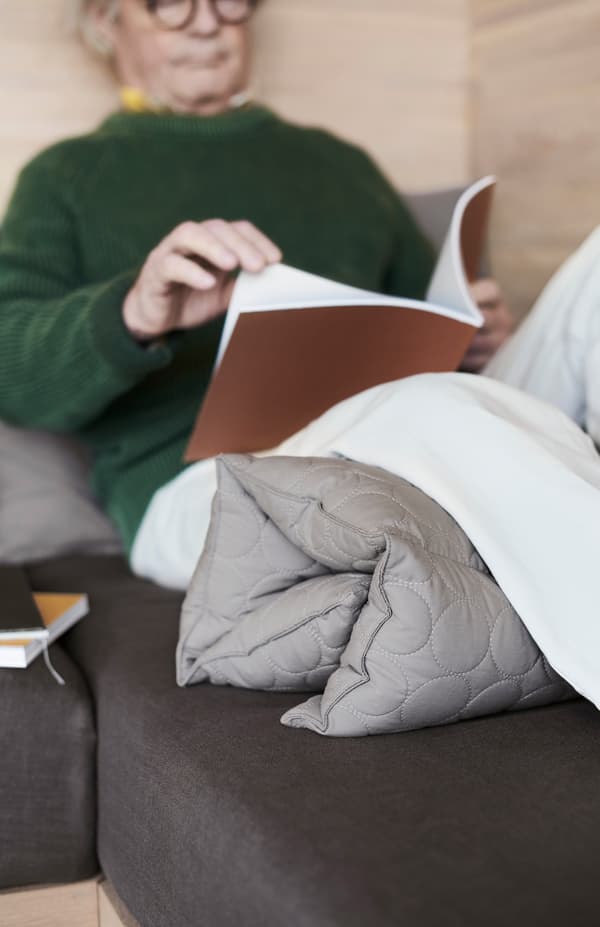 A man in his 60s leans back against the wall reading. OMTÄNKSAM multi cushion is tucked under his knees.