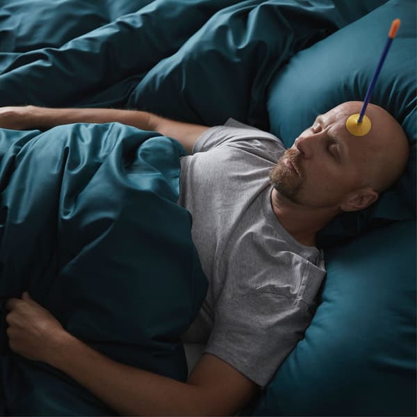 A man lies asleep in a plush-looking bed, with a toy suction-cup arrow stuck to his forehead.