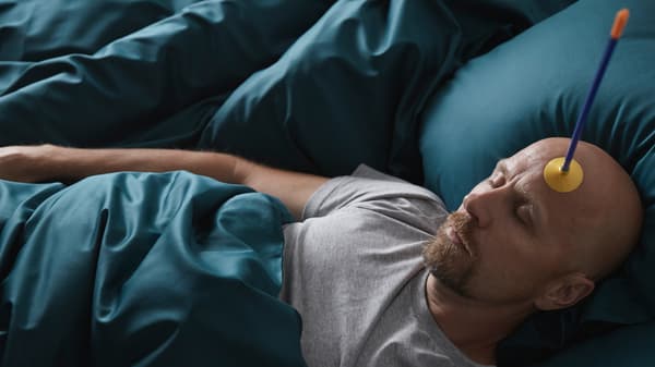 A man lies asleep in a plush-looking bed, with LUKTJASMIN bed linen. He has a toy suction-cup arrow stuck to his forehead.