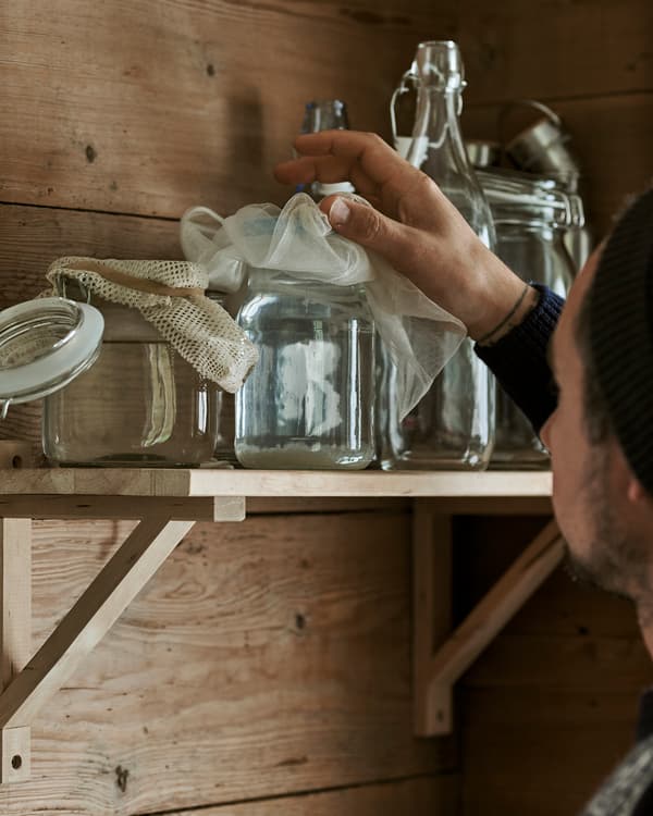 A man picks up a glass Kilner-style jar with muslin cloth lid from a wooden shelf.