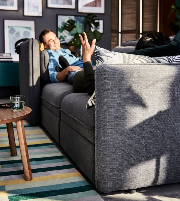 A man relaxing on a grey VALLENTUNA modular sofa, placed on a multi-coloured rug in a living room setting.
