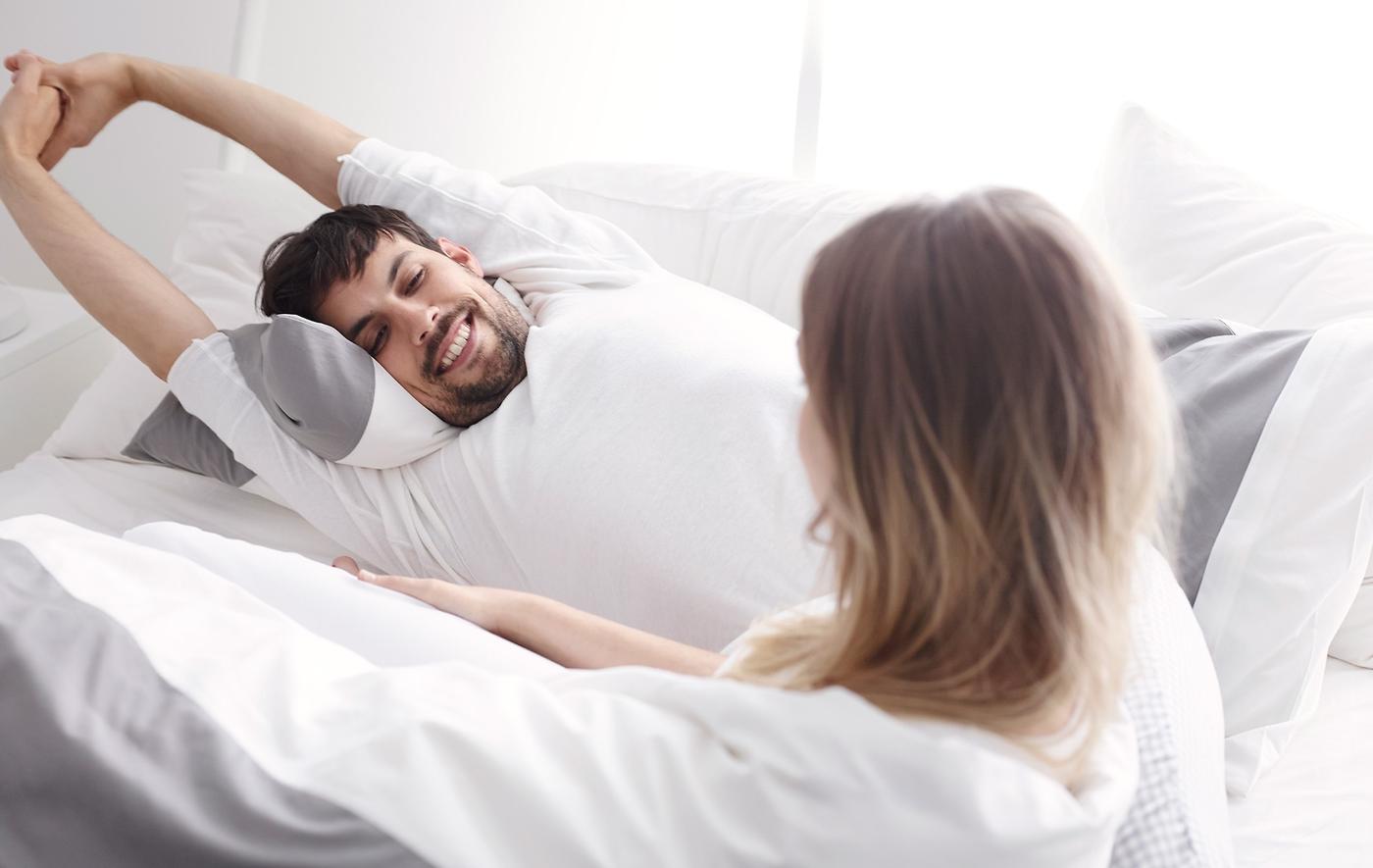 A man stretching his arms in bed whilst he talks to a woman.