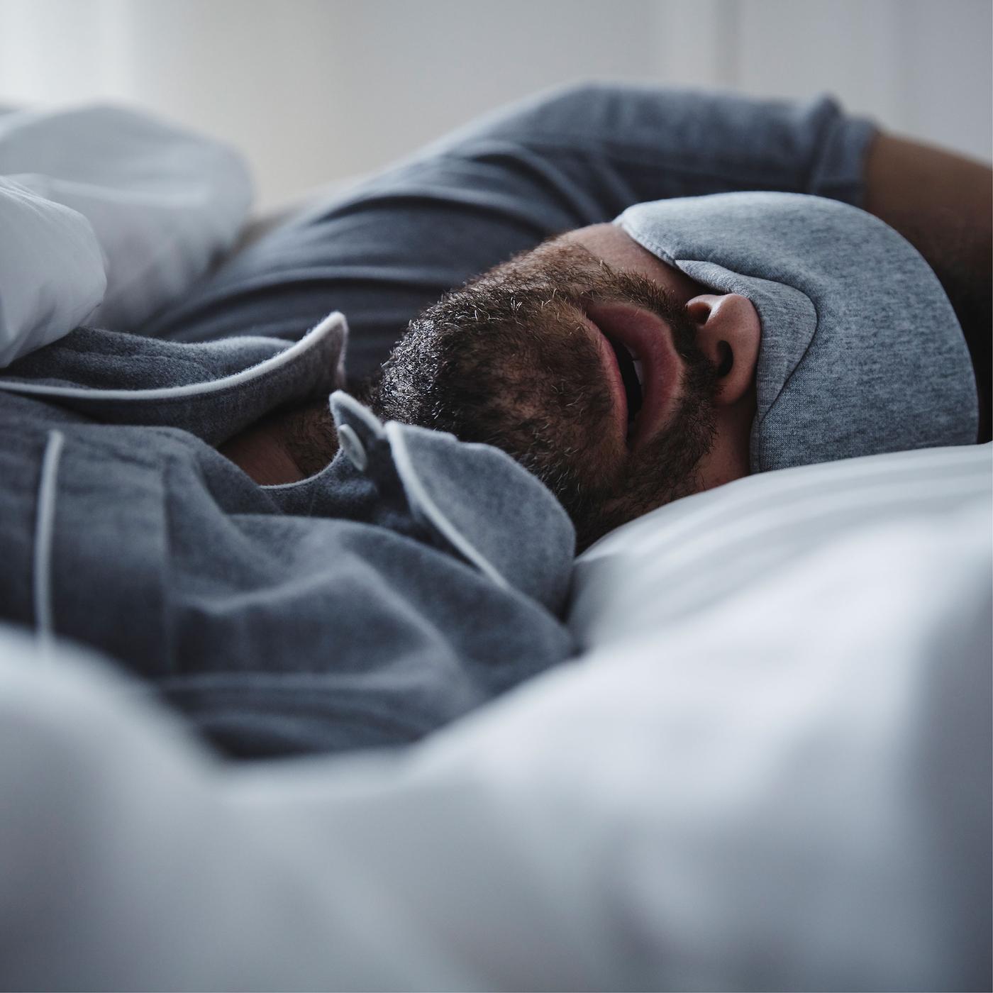 A man wearing blue pyjamas and matching eye-mask lies asleep in bed with his mouth open.