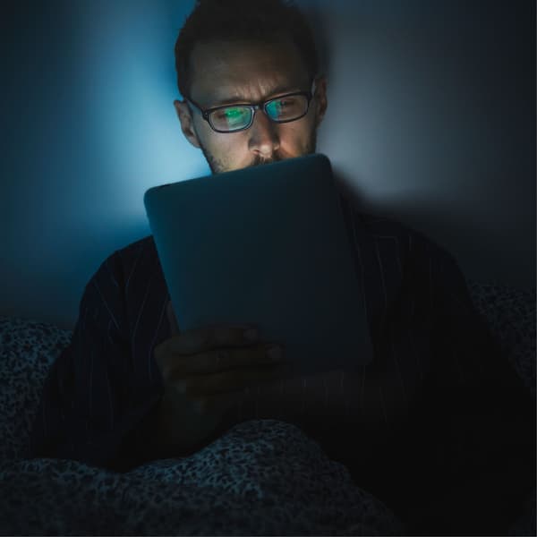A man wearing glasses looks at an iPad in bed, its light reflecting on his tired-looking face.