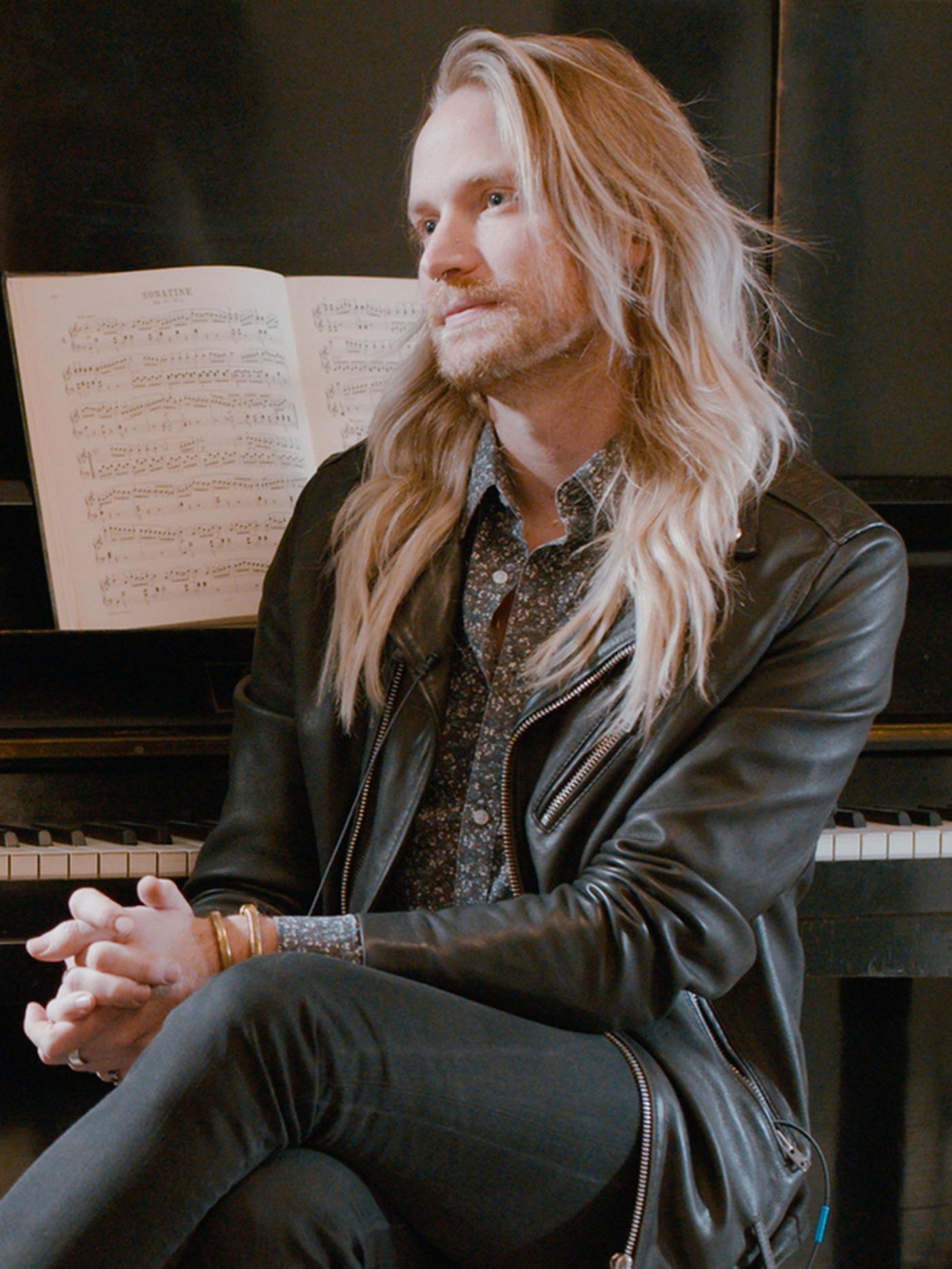 A man with a blonde beard and blonde long hair, sitting on a stool by a piano.