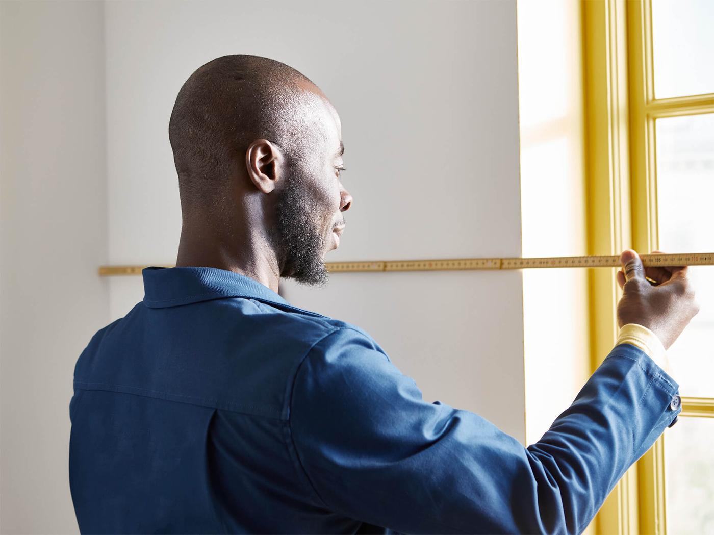 A man with a blue jacket is measuring a wall next to the window.