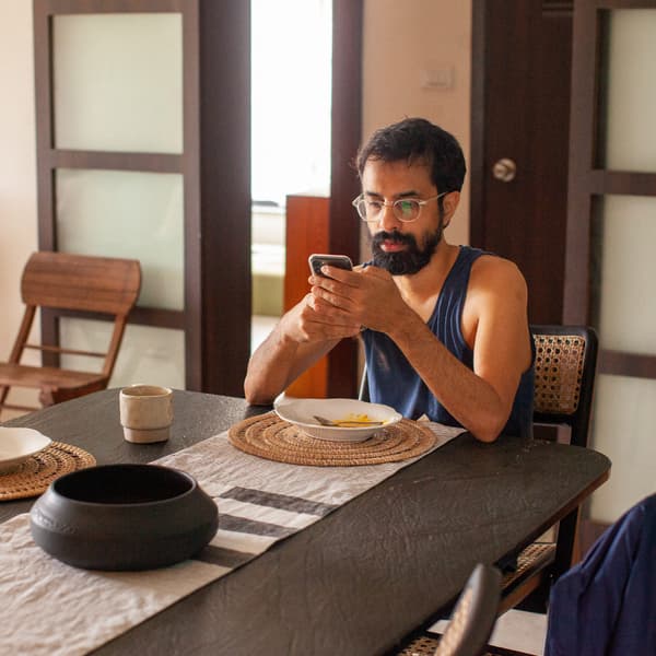 A man with dark hair and a beard just finished eating and is browsing his mobile phone at the kitchen table.