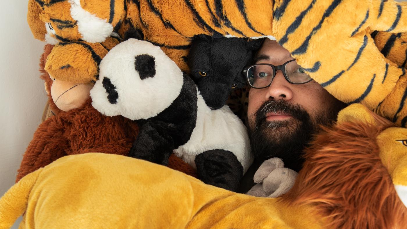 A man with glasses and a beard lies among a whole bunch of DJUNGELSKOG soft toys. Only his face shows.