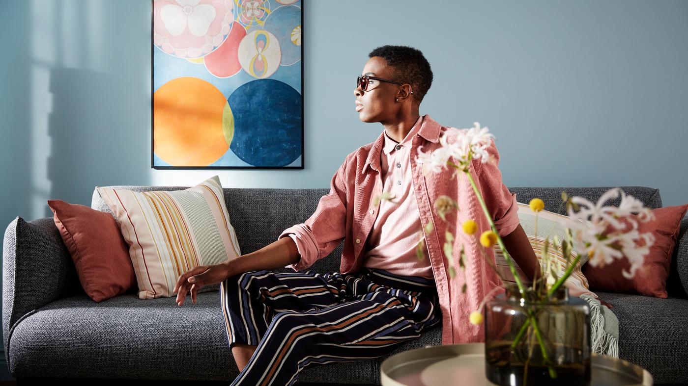 A man with pink and striped clothes sitting in a grey sofa placed against a wall with a large poster of pastel art.