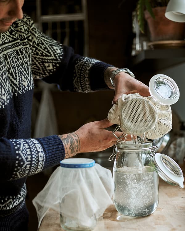 A man works at a kitchen bench, pouring clear liquid from one Kilner-style jar to another using muslin as a strainer.