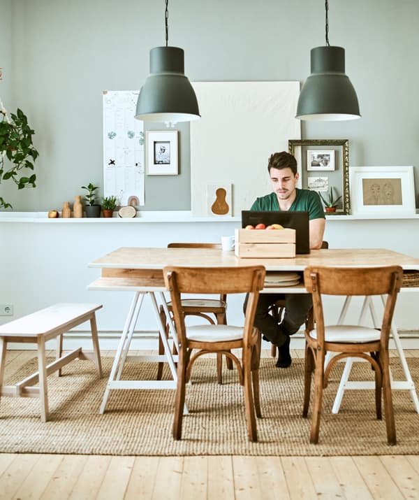 A man works on a laptop at a wood table, two grey pendant lamps hang above with a display of pictures on a grey wall behind.