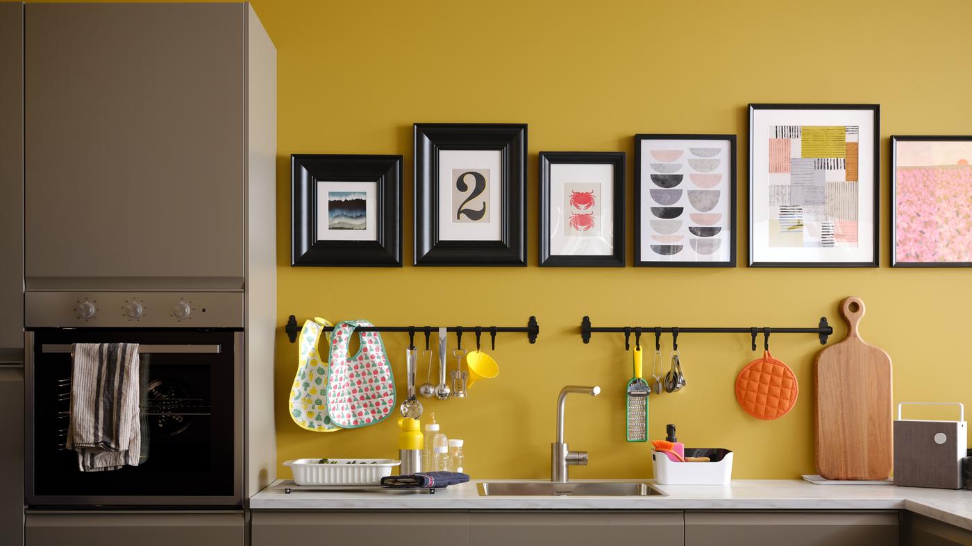 A matt dark beige kitchen with a white marble effect worktop, plus black rails and pictures in black frames on a yellow wall.
