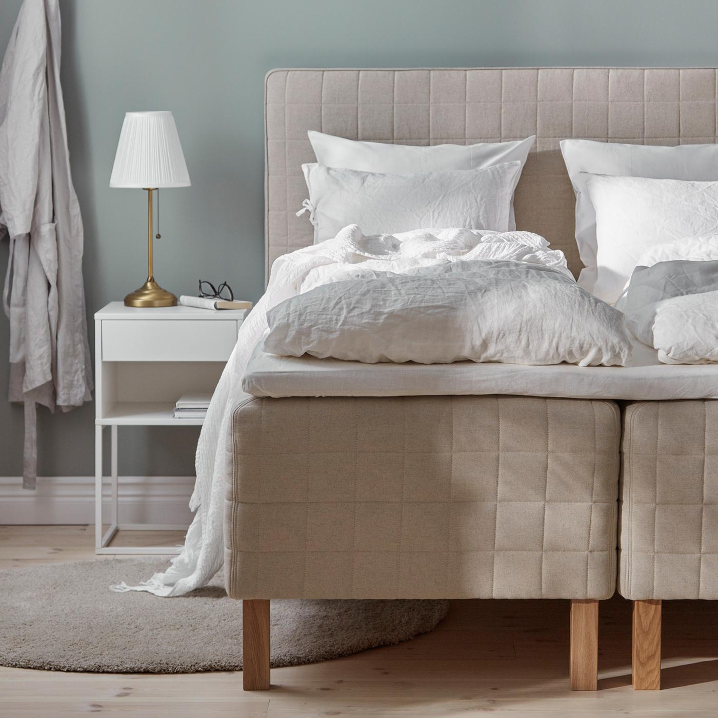 A mattress with headboard stands in a bedroom with a round rug. A bathrobe hangs from a hook on the wall nearby.