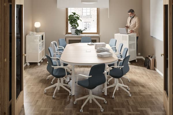 A meeting room with a conference table and 10 chairs. A man stands at the back by a window, behind a computer on a cabinet.