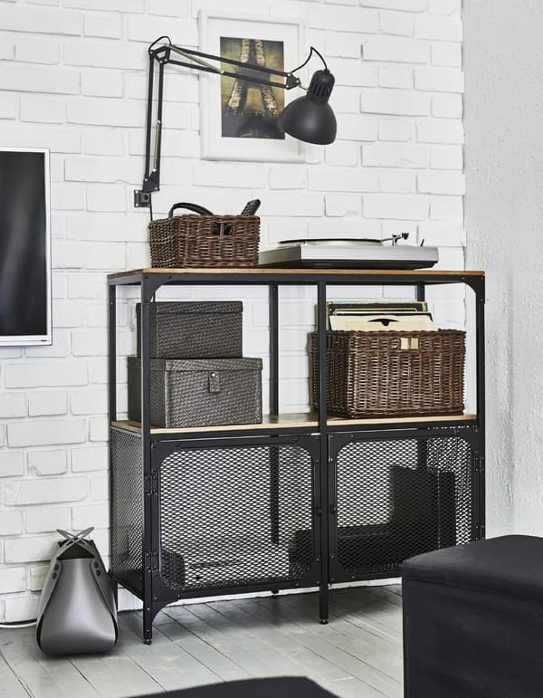 A metal FJÄLLBÖ shelving unit stores entertainment equipment like remotes and cables in a living room.