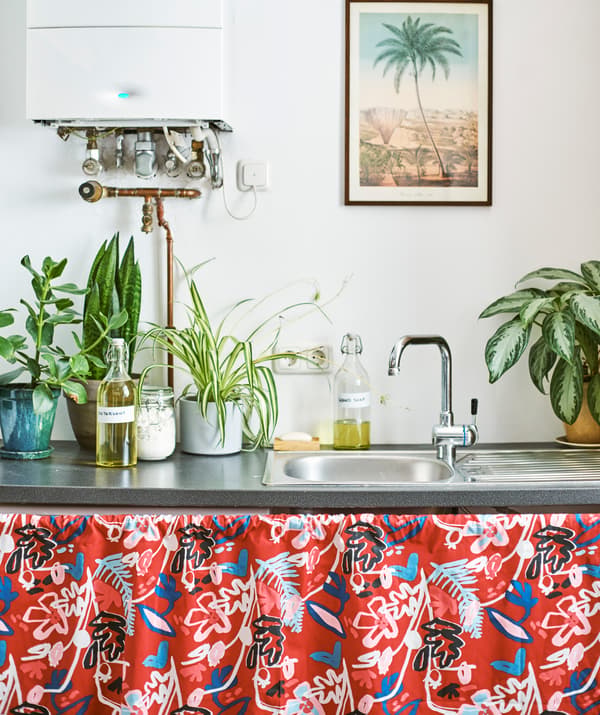 A metal sink with plants and bottles filled with liquid on top. Red patterned fabric covers under the sink.