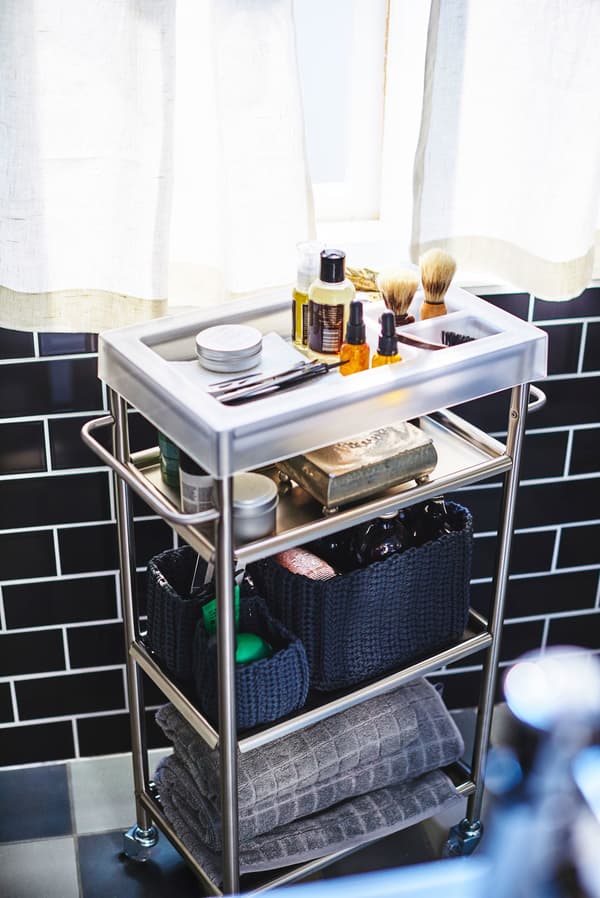 A metal trolley in a black tiled bathroom stores mens grooming products, like shaving equipment and towels.