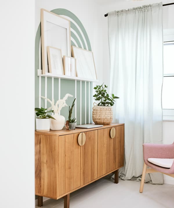 A mid-century modern style sideboard by a wall with a painted green arch motif and picture ledge with framed artwork.