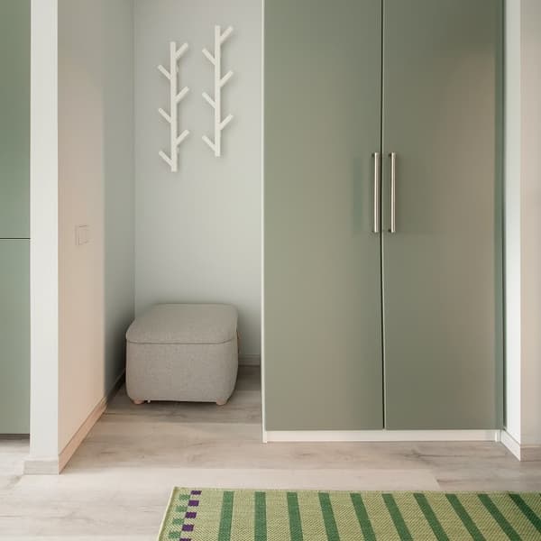 A minimalist hallway corner featuring a pair of sleek sage green wardrobes with vertical silver handles. To the left, a cozy light gray ottoman sits neatly in a recessed nook under two modern white wall hooks shaped like branches. The flooring is a light wood laminate, and part of a green woven rug with purple and dark green stripes adds a touch of color and texture to the space. The overall look is clean, modern, and functional.