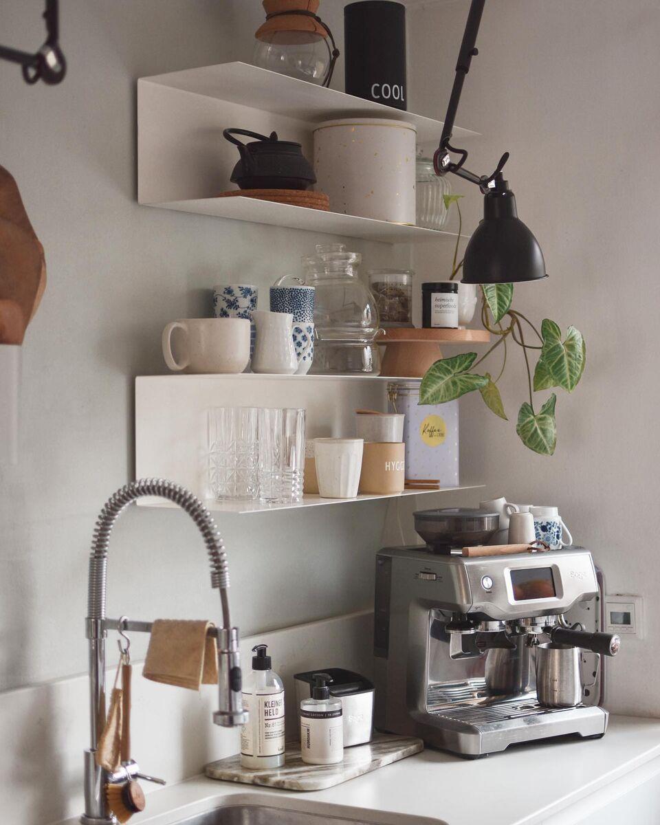A minimalistic coffee corner in the kitchen, featuring an espresso machine on the countertop, white wall shelves with cups and glassware, a black wall lamp, a green plant, and a stainless steel tap.