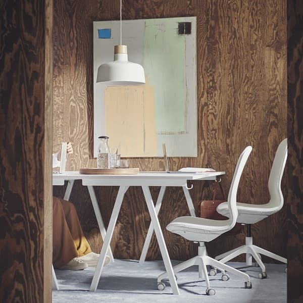 A modern and stylish small meeting or workspace featuring a white table with angled legs and two ergonomic office chairs. The room is surrounded by dark wood panel walls with a prominent natural grain texture. A minimalistic pendant lamp hangs above the table, which holds a glass water carafe, cups, and a wooden tray. A laptop and notebook rest on the table. Abstract art with muted tones of green, beige, and black hangs on the wall, adding a touch of color. A person partially visible is seated at the table, wearing mustard-colored pants and white sneakers. The overall mood is calm, focused, and creative.
