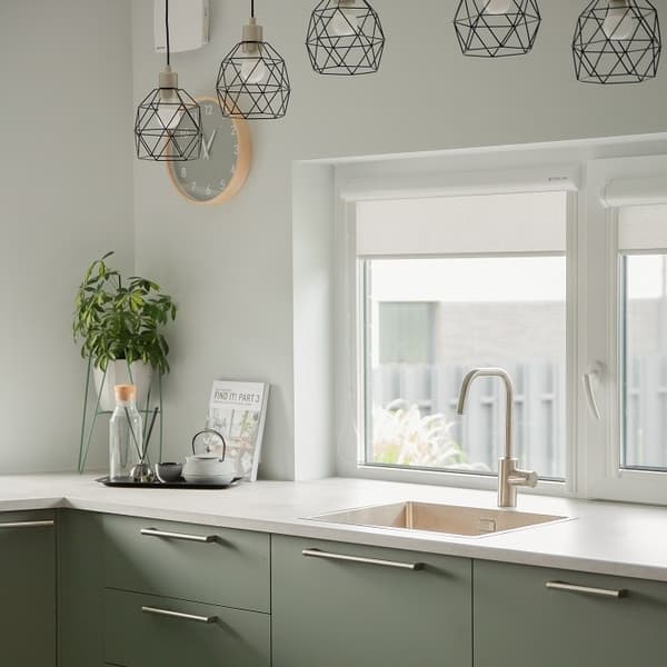 A modern kitchen corner with soft sage green lower cabinets and a light beige countertop. A stainless steel faucet is centered beneath a large double window, which lets in ample natural light. Above the counter hang stylish black geometric pendant lights, adding a contemporary touch. On the left side of the countertop sits a black tray with a glass water carafe, teapot, and a tall green plant in a glass vase. A round wall clock with a wooden frame adds a natural accent, while a cookbook titled 