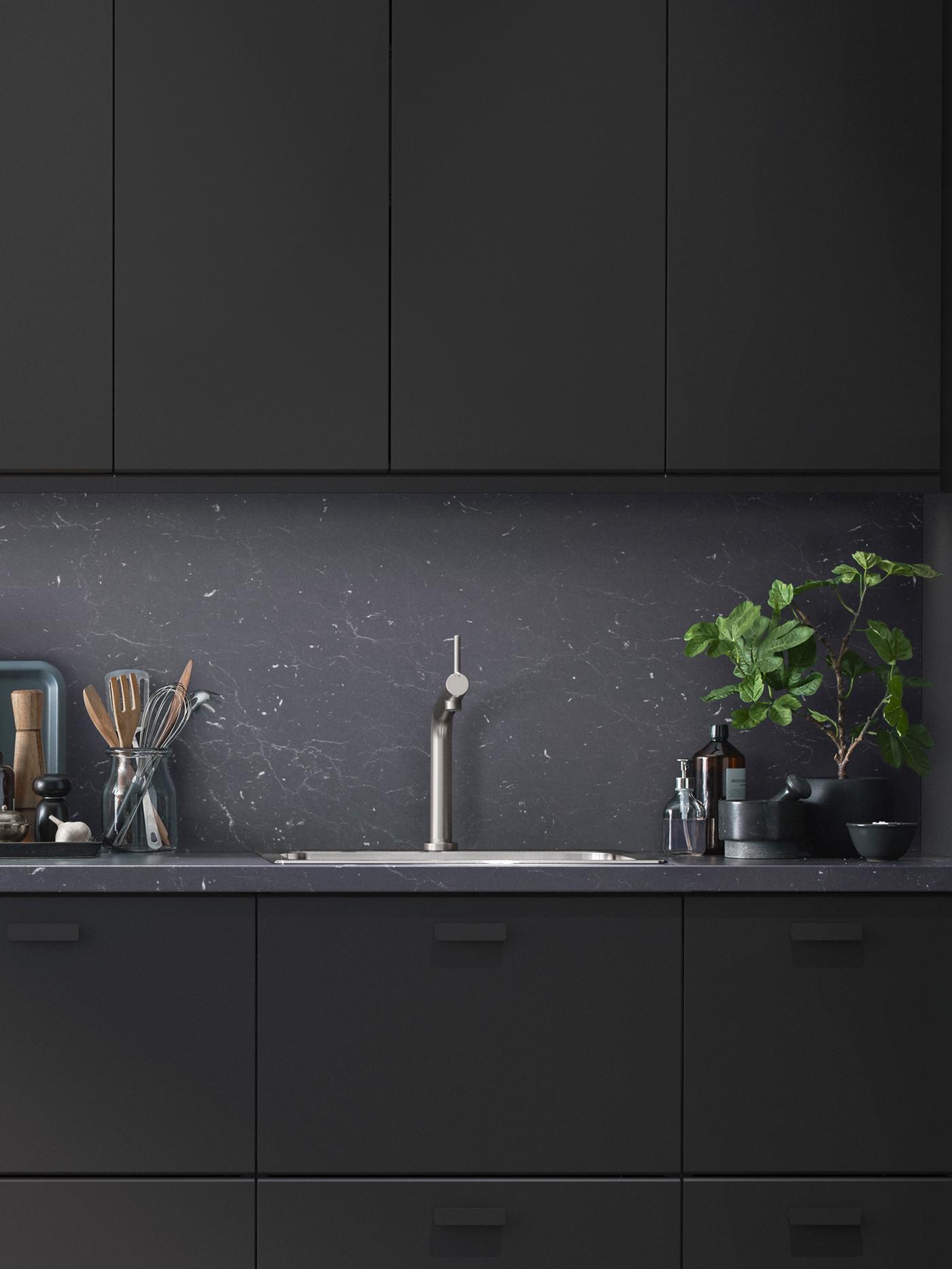 A modern kitchen with black KUNGSBACKA fronts and utensils on one side of the sink and a large plant on the other.