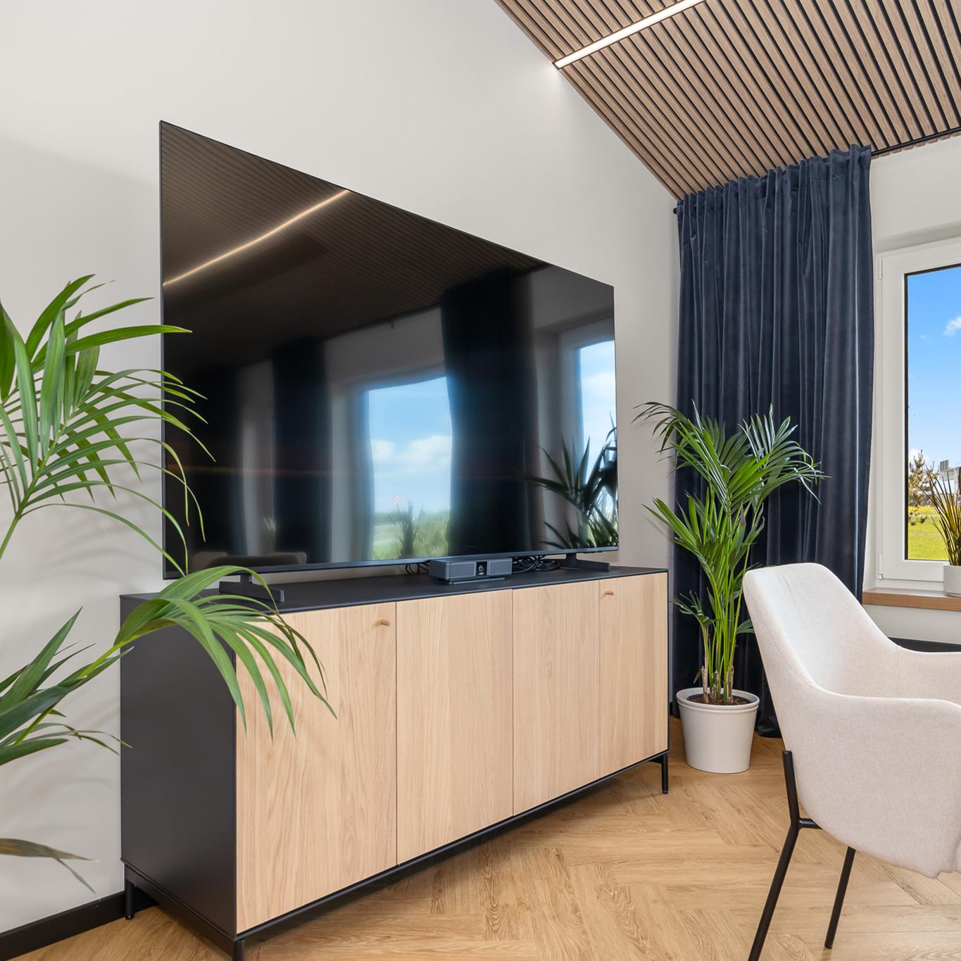 A modern lounge-style meeting room corner featuring a large flat-screen TV mounted above a light wood cabinet, surrounded by tall potted plants and soft natural light from a nearby window. The space includes a cushioned chair, dark curtains, and warm wood flooring.