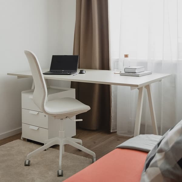 A modern minimalist workspace featuring a white desk with a matching white office chair on wheels. A laptop, pen, and mouse sit on the desk, alongside a couple of books, a clear glass, and a cork-topped carafe. The setup is placed in front of a window with sheer white curtains and brown blackout drapes, allowing soft natural light into the room. A beige rug lies under the desk, and part of an orange sofa with a gray cushion is visible in the foreground.