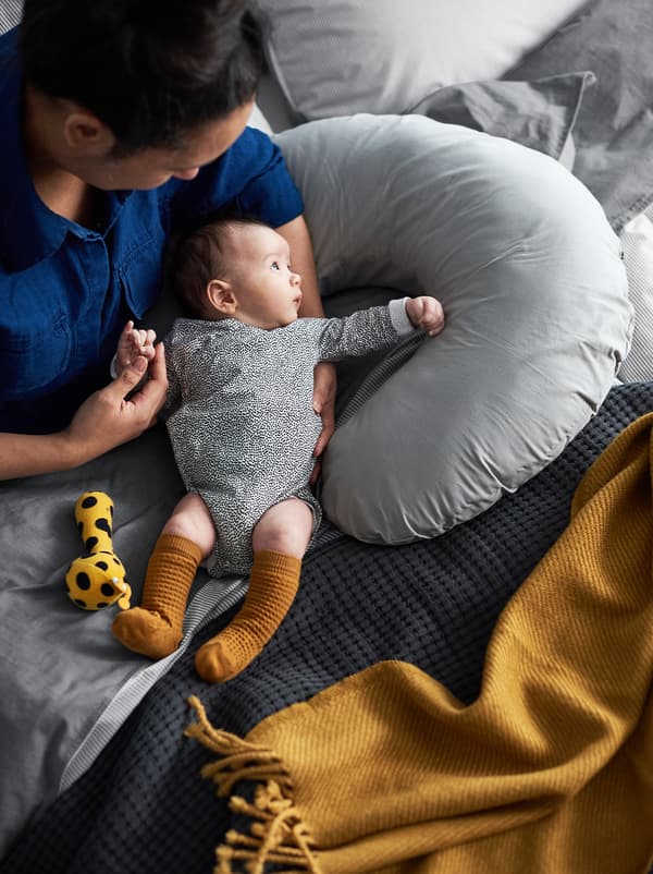 A mother and her baby sitting in a bed with covers in grey and mustard shades, and a nursing pillow beside them.