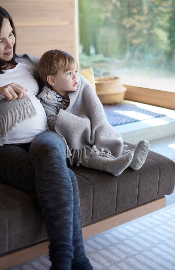 A mother and young child wrapped in a grey OMTÄNKSAM wool throw sit close together on a sofa.