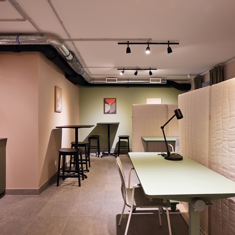 A multifunctional co-working space with shared desks, acoustic divider panels, a kitchenette, and lounge areas in the background. Industrial-style lighting and warm neutral tones create a welcoming atmosphere for work and collaboration.