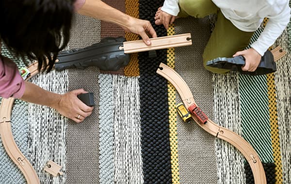 A mum and son build a wood road track together on a rug woven with different coloured stripes.