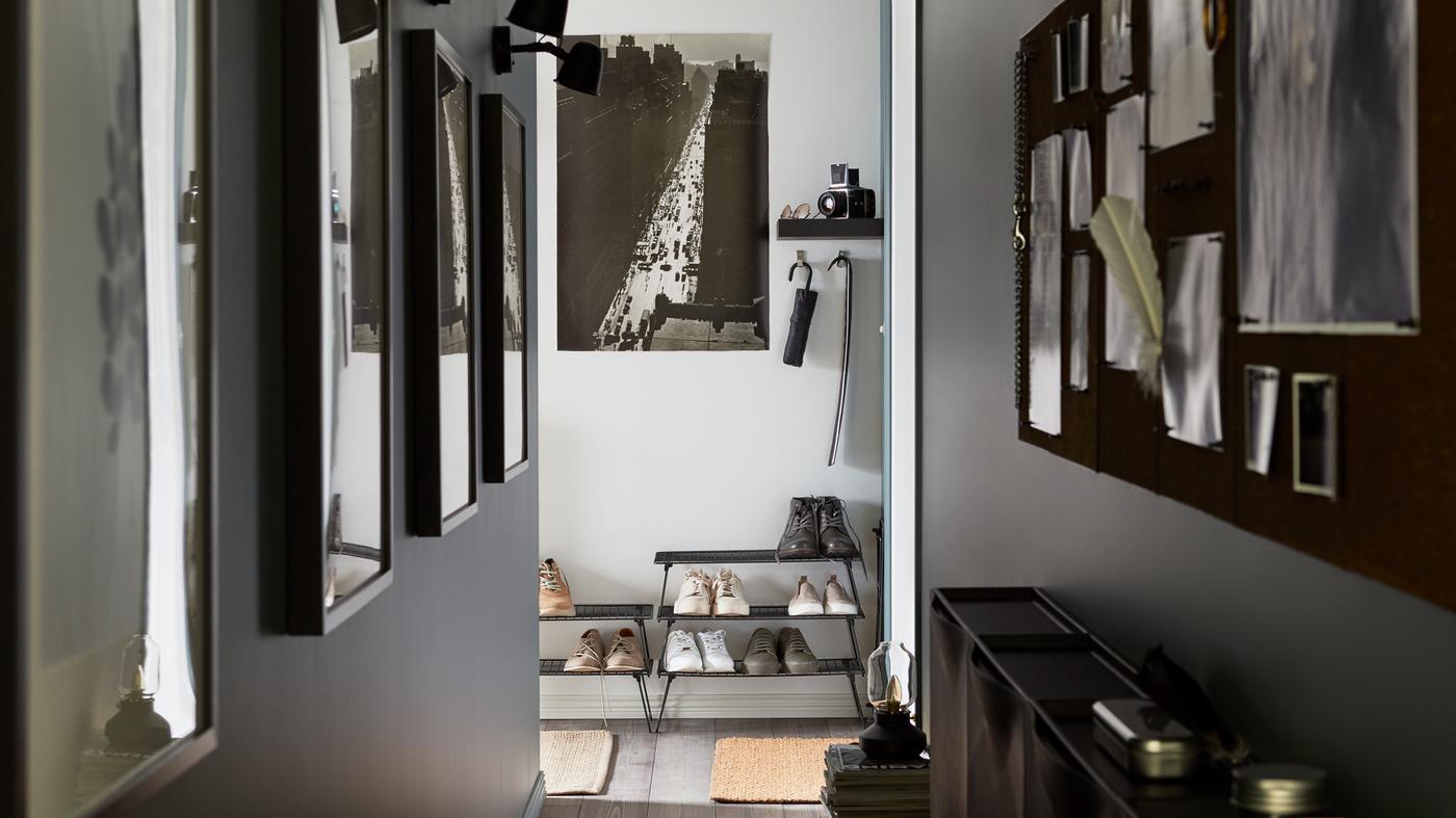 A narrow hallway with photos on the walls, black wired shoe storage stacked on top of each other, a black noticeboard.