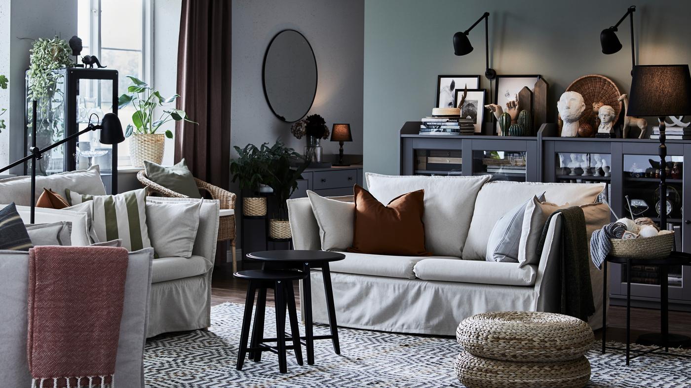 A natural BACKSÄLEN two-seat sofa with two black coffee tables in front of it and two grey HAUGA glass door cabinets behind.