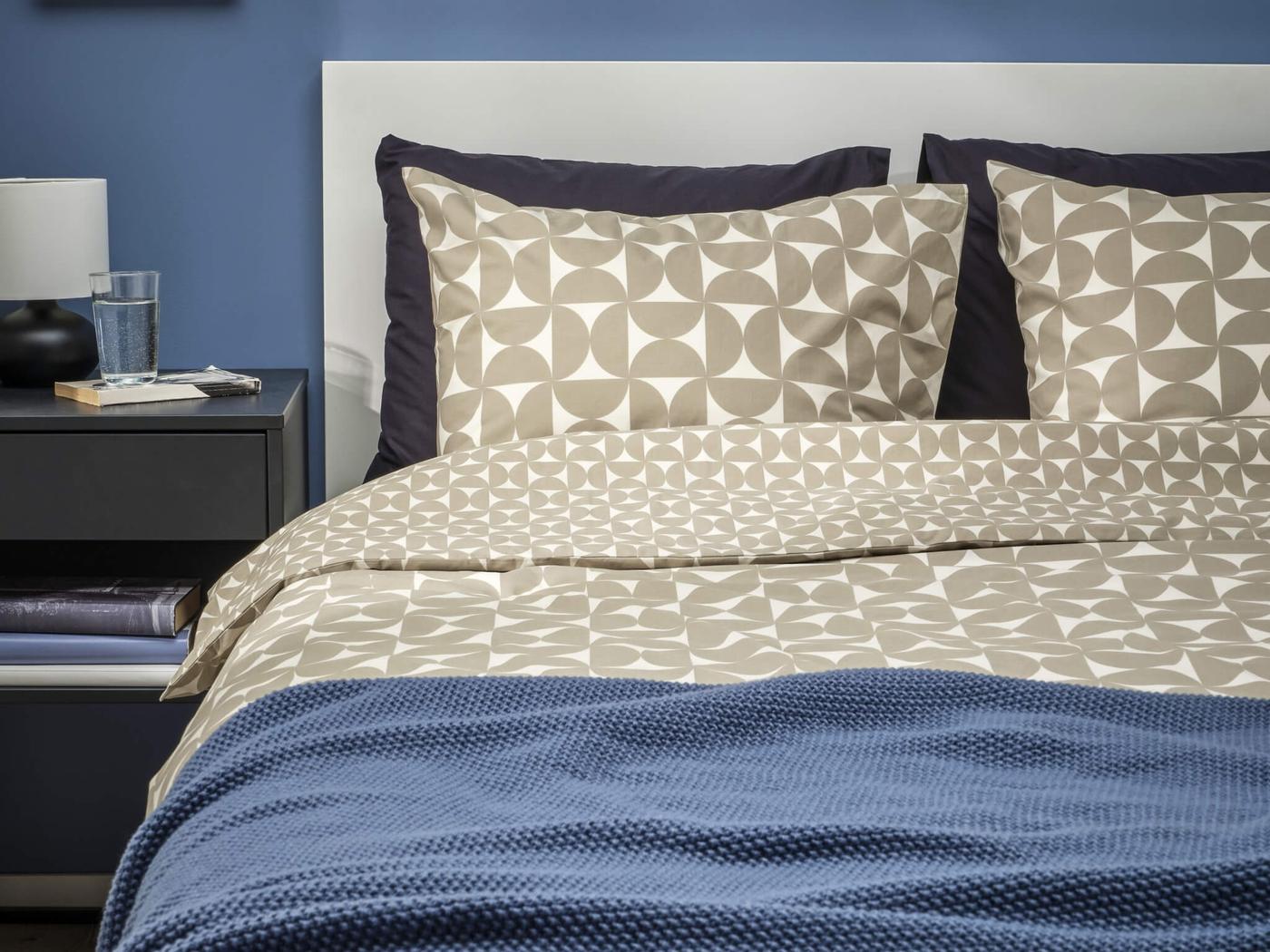 A neatly made bed with patterned pillows and a blue comforter in a bedroom with blue walls and a bedside table.