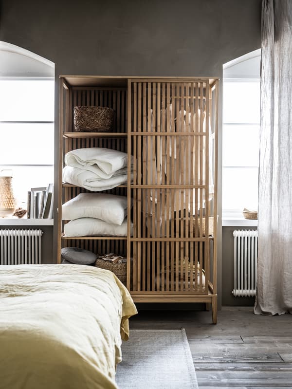 A NORDKISA open wardrobe filled with duvets, pillows, decorations and clothes stands in-between two bedroom windows.