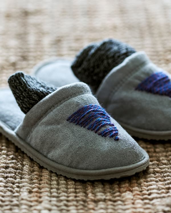 A pair of grey slippers on a jute rug, customised with blue woollen thread sewn into a fir tree motif on the front.