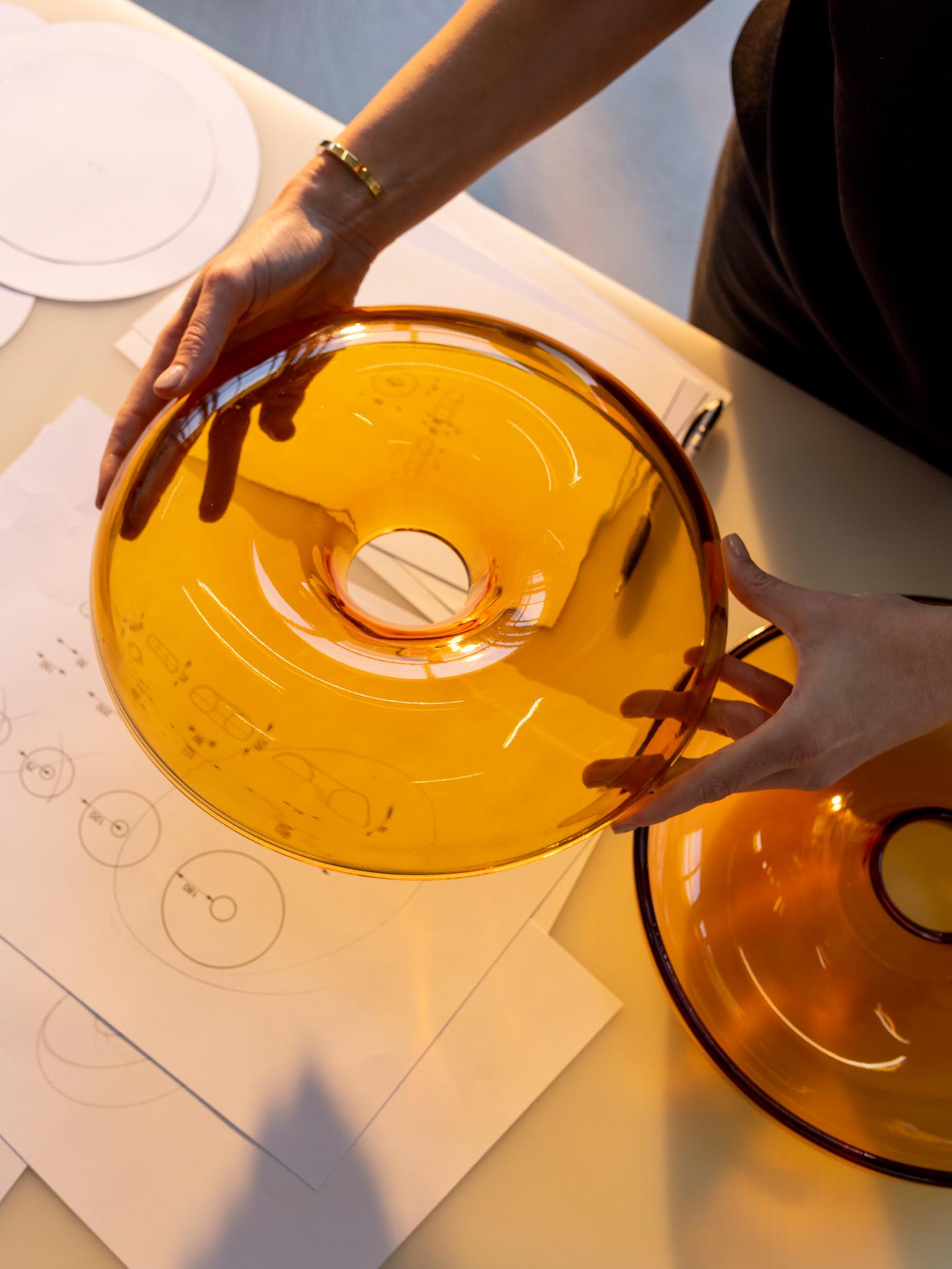 A pair of hands holds up a lid of an orange VARMBLIXT glass doughnut-shaped bowl above a table with sketches of the object.