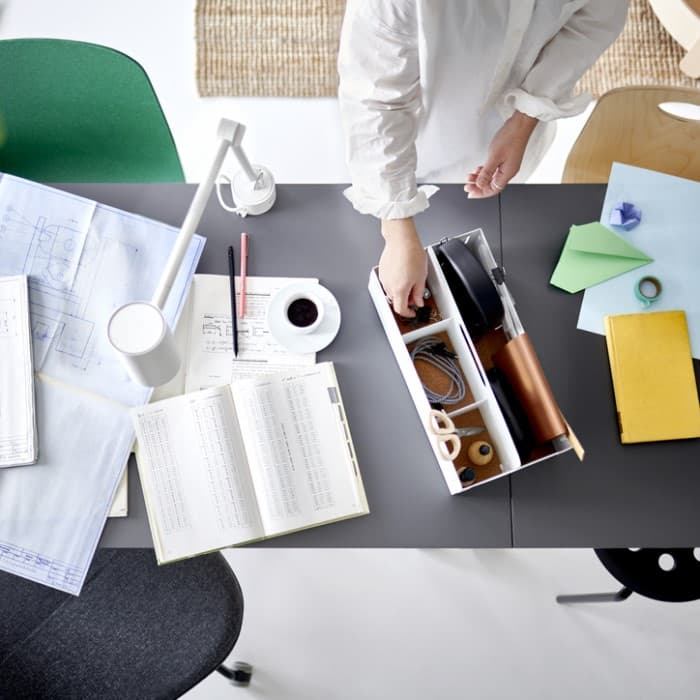 A pair of hands picking an item from a white desk organiser on two joined table tops with work papers and a white work lamp.