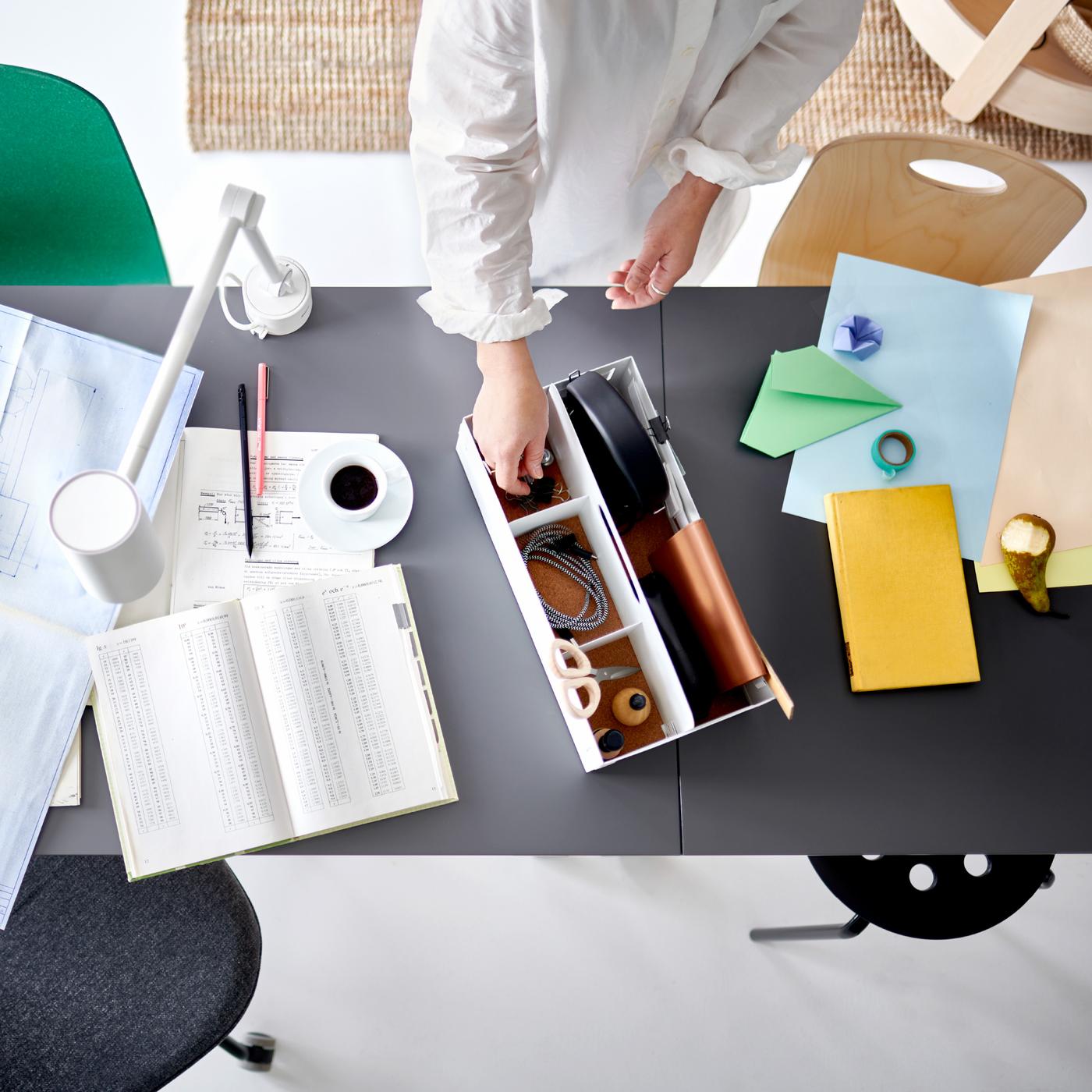 A pair of hands picking an item from a white desk organizer on two joined table tops with work papers and a white work lamp.