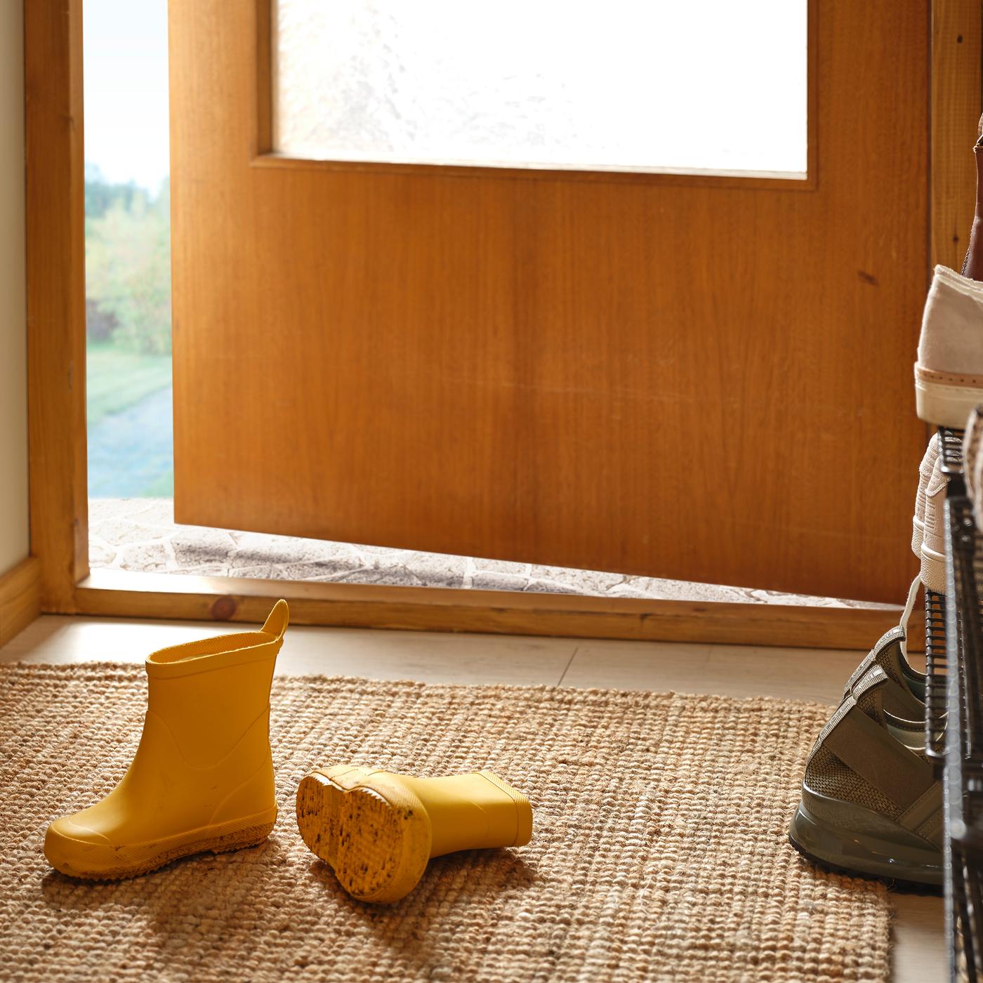A pair of yellow children’s boots lie on a LOHALS flatwoven rug in a hallway next to a partly open front door.