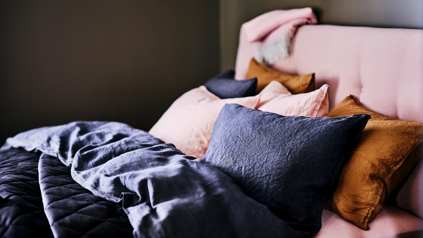 A pale pink IDANÄS upholstered bed with storage, covered in a dark grey duvet and pillows with pale pink sheets.
