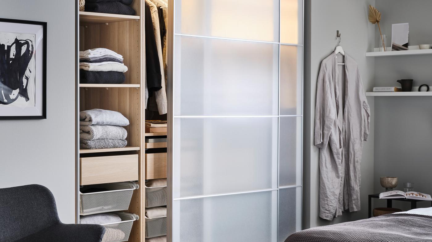 A partly open PAX/SVARTISDAL wardrobe with shelves, drawers and hanging clothes stands in a bedroom near a bed.