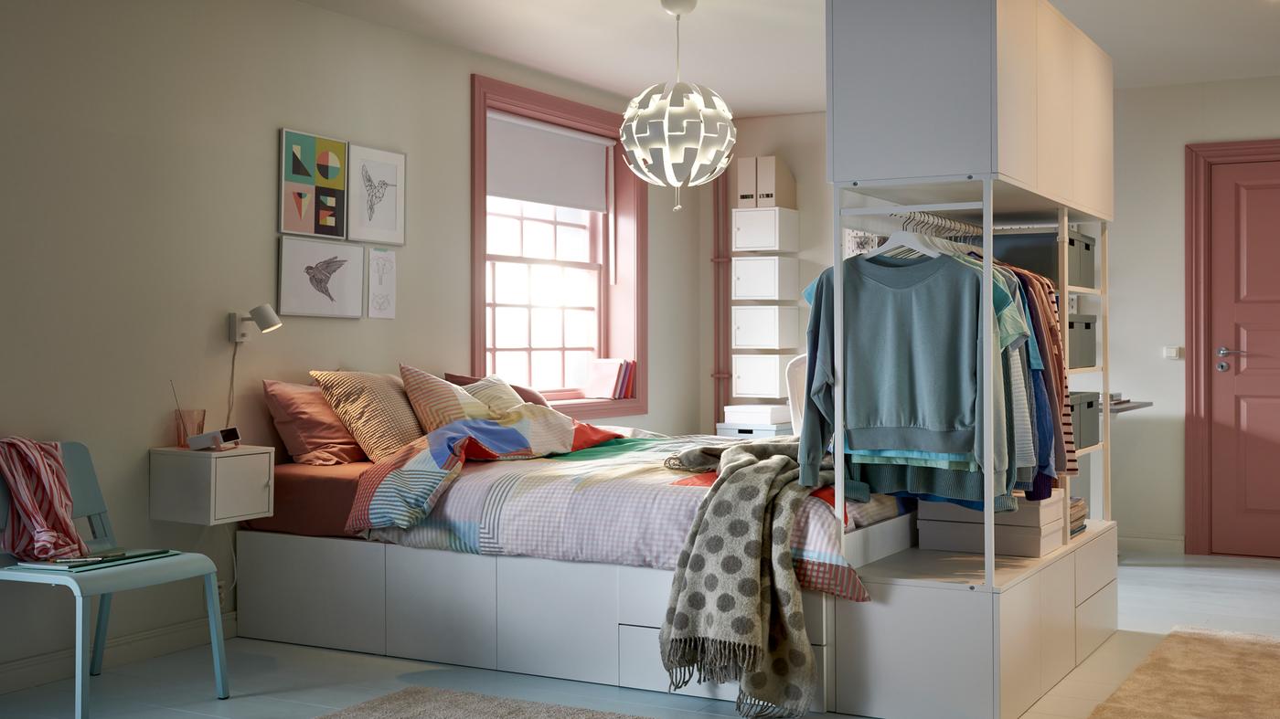 A pastel student room with a bed in focus with integrated storage underneath and hanging clothes storage at the foot end.